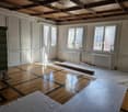 Empty room with wooden flooring, large windows with white curtains, and a fireplace on the left side.