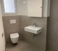 Modern bathroom with a toilet, wall-mounted sink, chrome faucet, and mirrored cabinet with built-in lighting. The walls are tiled in a neutral color scheme, and the floor is covered with beige tiles.
