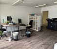 Office space with wooden flooring, two desks, chairs, multiple computer screens, keyboards, a printer, shelves with boxes, and a small plant.