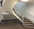 Spiral staircase with dark stone steps and a modern metal railing, leading up to a higher level. The hallway has a dark, textured floor and white walls with a curved ceiling.