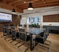 conference room with a large table, chairs, projector screen, wooden floor, wooden ceiling, windows, whiteboard, plant, and a cabinet with fruits
