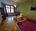 Bedroom. Cozy bedroom with wooden flooring, large windows with purple curtains, a bed with a red checkered duvet, a white armchair, a small table, a wardrobe, and a potted plant.