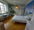 Bedroom. Cozy bedroom with wooden flooring, large windows, a double bed with white bedding, a desk with a chair, a cabinet with mirrored doors, and decorative plants.