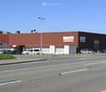 A large industrial-style building with a red metal roof and signage for "FIRSTCARS" visible. The building has multiple entrances and a parking area with several cars.