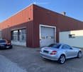 A large industrial-style building with a red metal facade, large garage door, and several windows. Several cars are parked in front of the building on a paved area.