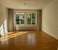 Empty room with wooden flooring, large windows with curtains, radiator, and natural light streaming in.