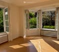 Empty room with wooden flooring, large windows, radiators, and natural light from the garden view.