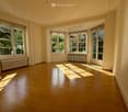 Empty room with wooden flooring, large windows with white frames, and radiators beneath the windows. Sunlight streams in, casting shadows on the floor.
