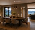 Elegant dining area with a large wooden table, six chairs, a vase with flowers, and candles. A bar counter with stools is visible in the background, with expansive mountain and lake views through large windows.