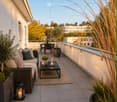 A modern balcony with a sofa, coffee table, dining set, and decorative lanterns, surrounded by potted plants and a view of trees and buildings.