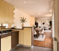 Open-plan kitchen and living area with light wood cabinets, granite countertops, a modern sink, a dishwasher, a white sofa, a coffee table, dining chairs, a dining table, decorative vases, and a potted plant.
