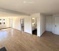 Empty room with wooden floors, white walls, large windows, and a radiator. There are doors leading to a bathroom and a bedroom.