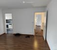 Empty room with wooden flooring, white walls, and open doors leading to a kitchen and another room. No furniture or equipment is present.