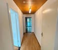 A long, narrow hallway with light wooden flooring, white walls, and a wooden ceiling. The hallway features several doors and a light fixture hanging from the ceiling.