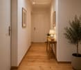 Modern hallway with light wooden flooring, white walls, and subtle lighting. Features a small wooden console table with a lamp, bowl, and potted plant, along with framed artwork on the walls.