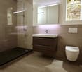 Modern bathroom with a glass-enclosed shower, a dark wooden vanity with a white sink, a toilet, and a large mirrored cabinet above the sink. The floor is tiled in a warm beige color.