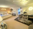 Elegant bedroom with a large bed, fireplace, nightstand, and a sitting area with chairs and a table. The room features modern lighting and a cozy ambiance.