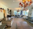 A spacious, well-lit living and dining area with a beige sofa, coffee table, dining table with chairs, and decorative lamps. The room features large windows and a chandelier, creating a bright and inviting atmosphere.