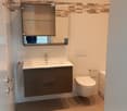 Modern bathroom with a white sink, dark grey cabinet, toilet, and a large mirror with built-in storage.