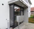 Modern building entrance with glass door, mailboxes, and small garden area with wooden fence.