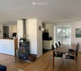 Open-plan living and dining area with wooden flooring, a central wood-burning stove, a dining table with chairs, a kitchen area with cabinets and appliances, and a large window with blinds.
