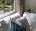 A cozy living room corner with a white sofa, multiple pillows, a blue blanket, and a decorative elephant statue on a windowsill, overlooking a serene garden with a pond and water fountain.