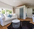 Bright, modern living space with a white sofa, coffee table, and a small kitchen area featuring white cabinets and a microwave. The room has wooden flooring, large windows with curtains, and a black area rug.