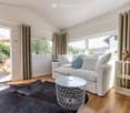 Bright living room with white sofa, coffee table, side table, and a large window with curtains. The room features wooden flooring and a dark rug.
