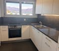 Modern kitchen with white cabinets, dark countertops, stainless steel oven, and a sink with a faucet. Large windows offer a scenic mountain view.