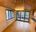 Empty room with wooden flooring, white walls, large windows with blinds, and a sliding glass door leading to a balcony area.