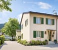 Two-story house with light beige walls, green shutters, and a well-maintained garden with colorful flowers and shrubs.