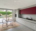 Modern kitchen with white and red cabinets, a black countertop, and a dining area with a round table and chairs. Large windows offer a view of a garden.