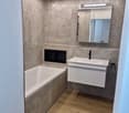 Modern bathroom with a bathtub, sink, and mirror. The walls are covered in textured grey tiles, and the floor is made of light wood. LED lighting runs along the ceiling edges, creating a sleek and contemporary look.