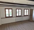 Empty room with wooden flooring, white walls, and three windows with wooden frames. A radiator is visible under the windows.