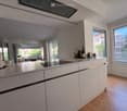 Modern kitchen with white cabinets, stainless steel countertop, induction stove, and large windows overlooking a garden area.