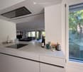 Modern kitchen with white cabinets, induction stove, and a large window overlooking greenery. Countertop has various items including a bottle, cup, and small appliances.
