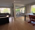 Living room with hardwood floors, large windows, and a balcony. Features a grey sectional sofa, a dining table with chairs, and a bowl on the table.
