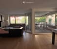 Bright living room with wooden flooring, large windows, and sliding glass doors leading to a balcony. Features a grey sectional sofa, a chair, a small table, and a television.