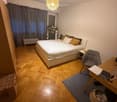 Cozy bedroom with wooden flooring, a large bed with pillows, a chair, a small table, a stool, and a hanging lamp.