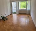 Empty room with wooden flooring, large windows with white curtains, and a small balcony visible through the open door.