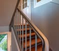 Modern staircase with wooden steps and a metal railing, leading to an upper level. The walls are painted in neutral tones, and there is a window at the bottom of the stairs offering a view of the garden.