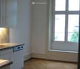 Empty kitchen with wooden flooring, white cabinets, a sink, and a dishwasher. Large windows allow natural light to enter.