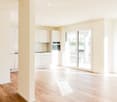 Bright, empty room with wooden flooring, white walls, and large glass sliding doors leading to an outdoor area. The room features a small kitchen area with white cabinets and a built-in oven, but no furniture or appliances are present.