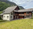 Mountain house with wooden exterior, a chimney, glass windows, and located in a valley with mountains in the background.