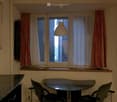 A small, furnished room with a glass table and two chairs, a window with curtains, and a hanging lamp.