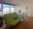 A modern living room with a green futon sofa, a small bookshelf, a toy, and picture frames on the wall. Large windows offer a scenic view.