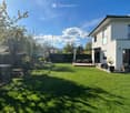 Well-maintained lawn with a modern house in the background, featuring a patio with outdoor furniture, a small garden area, and a swimming pool partially visible on the left side.