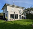 Two-story modern house with white exterior, large windows, and a spacious lawn. Features include a covered patio area with outdoor furniture, a small garden with a tree, and a balcony on the upper level.