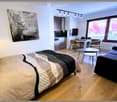A modern, open-plan living and sleeping area with a bed, sofa, coffee table, dining table, chairs, and a small kitchen area with a dishwasher and fridge.