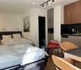 A well-lit, modern studio apartment with two single beds, a small kitchen area with cabinets and a dishwasher, a table with chairs, and a stool. The room features a painting on the wall and track lighting.